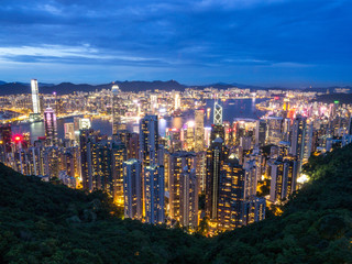 Hong Kong Skyline at Night from the Famous Victoria Point at Daytime. Green forest and river surrounded by towering sky scrapers which is densly populated. Deep blue sky and illuminated tall buildings