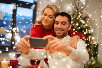 Obraz na płótnie Canvas christmas, holidays, technology and people concept - happy couple in taking selfie by smartphone at home dinner over snow