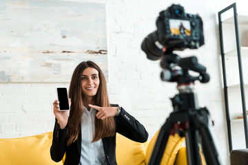Fototapeta na wymiar selective focus of happy video blogger pointing with finger at smartphone with blank screen near digital camera
