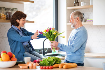 A senior couple in love indoors at home, man giving flowers to woman.