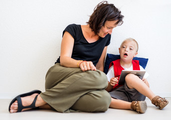 A down-syndrome school boy sitting on the floor with teacher, using tablet.