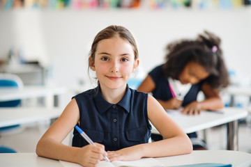 A small school girl sitting at the desk in classroom, looking at camera.