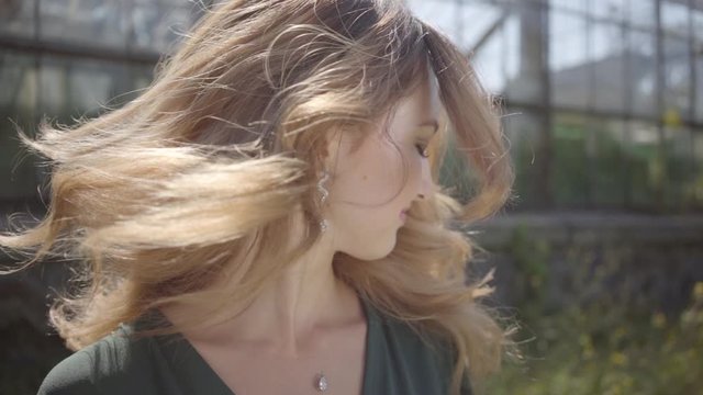 Portrait of cute young woman shaking her head with developing hair on a background of a greenhouse in the sun. Healthy leisure outdoors. Slow motion.