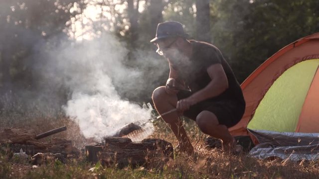 A young man in a hat fries grilled meat on a bonfire in the forest next to his tent. A lot of smoke from the fire. Series.