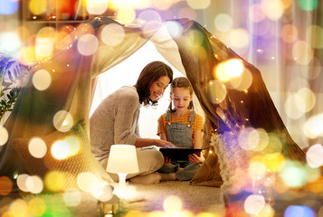 family, hygge and technology concept - happy mother and little daughter with tablet pc computer in kids tent at night at home