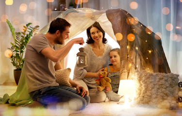 family, hygge and people concept - happy mother, father with lantern and little daughter with teddy bear playing in kids tent at night at home © Syda Productions