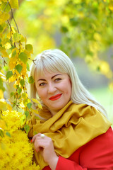 Portrait of a cute middle-aged blonde outdoors. She stands near a birch tree with yellow leaves in a red cloak with a bouquet of yellow flowers, looks at the camera and smiles.