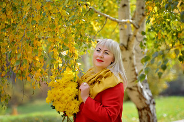 Middle-aged woman posing in the fall outdoors. She stands near a birch tree with yellow leaves in a red coat with a bouquet of yellow flowers and looks thoughtfully away. Fashion portrait. Close-up