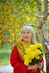 Large size woman with yellow flowers in autumn outdoors. She stands against a background of yellow-leaved trees in a red coat, looks away and smiles. Fashion portrait.