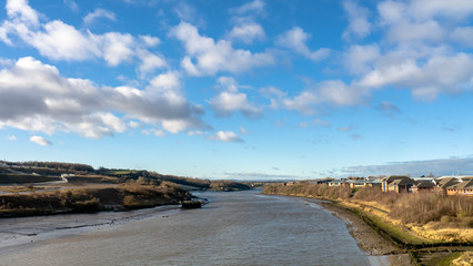 Landscape with blue sky and clouds taken from the Northern Spire Bridge with the river flowing towards the Tyne river in Newcastle. Also showing embankments either side and an old decayed boat.