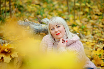 A bright middle-aged blonde poses on a Sunny autumn day in the forest in a gentle knitted jumper. Photo through the foliage. Woman's size plus xxl. Horizontal view