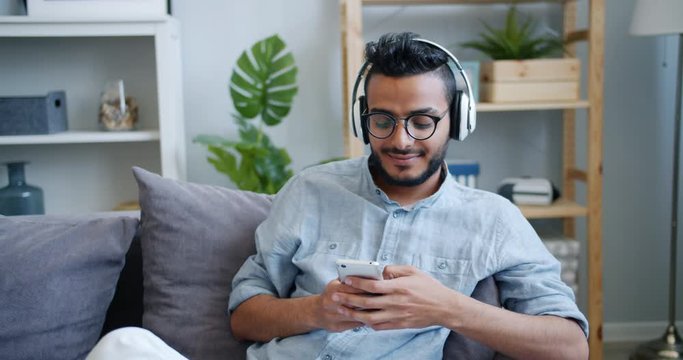 Handsome Arabian guy is using smartphone and listening to music in headphones sitting on couch at home. Modern lifestyle, devices and young people concept.