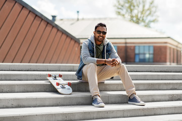 Obraz premium people and leisure concept - smiling indian man with skateboard sitting on stairs on city roof top