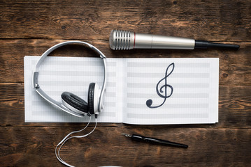 Music book, headphones, microphone and quill pen on a wooden table background.