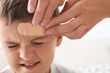 Woman applying adhesive bandage on boy's forehead indoors, closeup