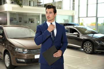 Young salesman with clipboard near car in modern dealership