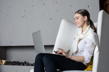 Young cheerful woman content manager or professional writer keyboarding on portable laptop computer, sitting in modern interior near fireplace background with copy space. Student learning online