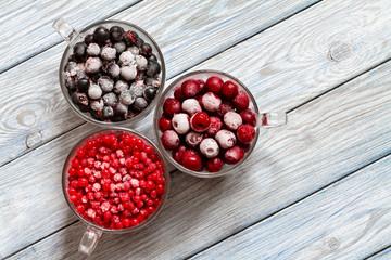 Ripe frozen sweet cherries, red currant and black currant with hoarfrost in the transparent glass cups on blue wooden background. Natural organic healthy food. Top view