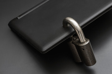 Locked steel padlock in a drilled hole of the black laptop on dark background. Concept of protecting personal data on a computer. A laptop is locked with a lock. Closeup, selective focus