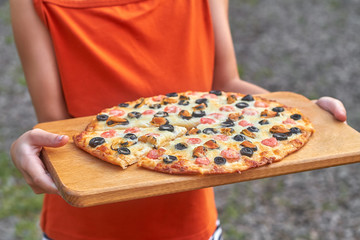 A girl in an orange t-shirt is holding homemade pizza on wooden board outdoors. Slice of pizza with shrimp, mussels, black olives and mozzarella. Home Holiday Cooking. Closeup, selective focus