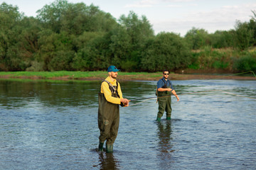 Two fishermen are standing in the river, wearing rubber boots. Men throw fishing rods. Fishing adventures