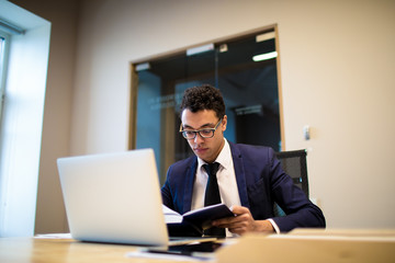 Professional Afro businessman in formal suit reading information in textbook, sitting at table with open laptop computer. Male entrepreneur preparing to meeting with staff, using netbook and notebook