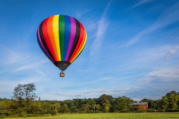 Rainbow colorful hot-air balloon floats over farm on a summer morning with bright blue sky 
