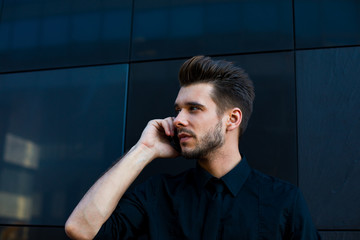 Young bearded businessman talking with partner via mobile phone, standing outside  company in work day. Male successful entrepreneur having smartphone conversation, looking away.Hipster guy calling