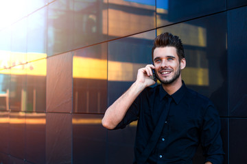 Happy smiling businessman dressed in formal wear talking with partner via mobile phone while standing outdoors against company near copy space for promotional content. Joyful entrepreneur calling