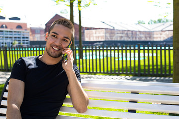 Fototapeta premium Young smiling hipster guy talking with friend via mobile phone while sitting on the bench in city park in beautiful sunny summer day. Cheerful man having pleasant cell telephone conversation