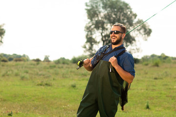 Happy fisherman in black glasses and a special suit poses in the nature. He holds his fishing rod...