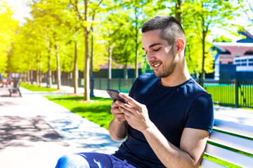Fototapeta premium Smiling hipster guy playing games in social network via mobile phone while relaxing in city park during free time, sitting near copy space for promotional text. Teenager with good mood using apps