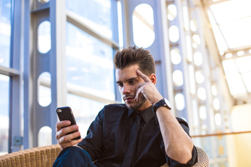 Confident businessman online booking via mobile phone, sitting in enterprise hall. Serious...