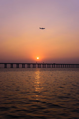 a vertical shot of sunset over the sea with an airplane on its way approaching to the airport