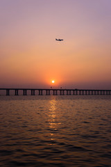 a vertical shot of sunset over the sea with an airplane on its way approaching to the airport