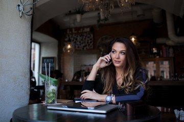 Young gorgeous woman talking via smartphone after distance work on laptop computer while sitting in modern coffee shop interior. Beautiful female having cellphone conversation with friend