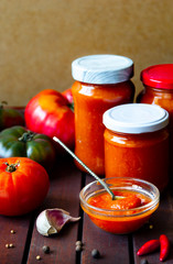 Homemade hot tomato sauce adjika in jars - Tomatoes, chilli pepper, garlic, herbs on wooden table.