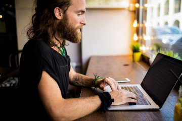 Fototapeta na wymiar Stylish hipster guy professional publication specialist typing promotional article for web page via laptop computer while sitting in coworking space. Bearded man content writer keyboarding on netbook