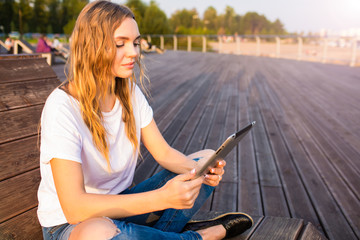 Female freelance social media content writer reading article on web site via touch pad while sitting outdoors in summer day. Woman university student reading electronic book on digital tablet