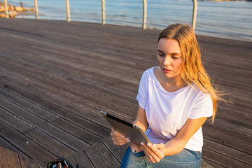 Gorgeous woman checking notification on touch pad while sitting outdoors near copy space for promotional content. Female blogger online banking via digital tablet during rest on sea quay in evening