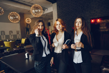 three beautiful elegant girls in white shirts and black jackets standing in a restaurant and use notebooks and pens, they also drink coffee and use the phone
