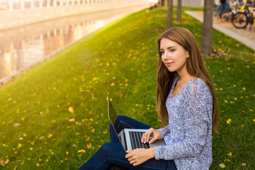 Attractive teenager girl with thoughtful look enjoying good day during distance work on laptop computer while sitting in beautiful autumn park near embankment river background with copy space