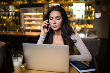 Fashionable attractive woman executive director talking via smartphone and reading financial information on laptop computer while sitting in restaurant during work break. Stylish female student 