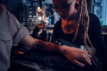 Young focused tattoo master is makining tattoo on client's hand at dark tattoo salon.