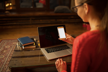 View from behind of a stylish hipster girl holding mobile phone with empty copy space screen background for promotional content while sitting with laptop computer at the table in home interior.