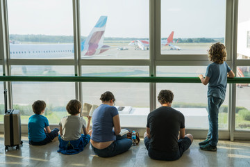 A large family is in front of the window at the airport. Four of them are sitting on the floor reading books, one boy is standing. Outside the window the plane is visible. Selective focus. Copy space.
