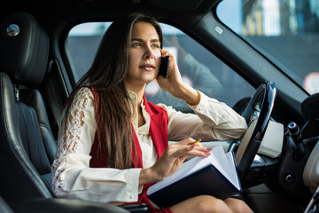 Confident woman executive director in formal wear talking via smartphone and using diary while sitting in automobile before work day in company. Female financier having cell telephone conversation
