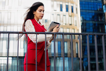 Confident woman professional manager reading e-mail on touch pad while standing outdoors against skyscraper building with copy space. Serious female employer using applications on digital tablet