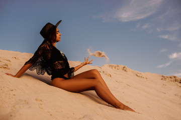 Outdoor full-length portrait of young beautiful woman with luxury tanned body wearing boho style crochet top, swimwear, hat, playing with sand, posing on beach, at sunset. Copy, empty space for text  © Victoria Fox