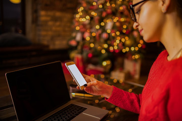 Woman blogger holding mobile phone with mock up empty copy space screen background with copy space for promotional content while sitting in home near Christmas tree during winter holidays